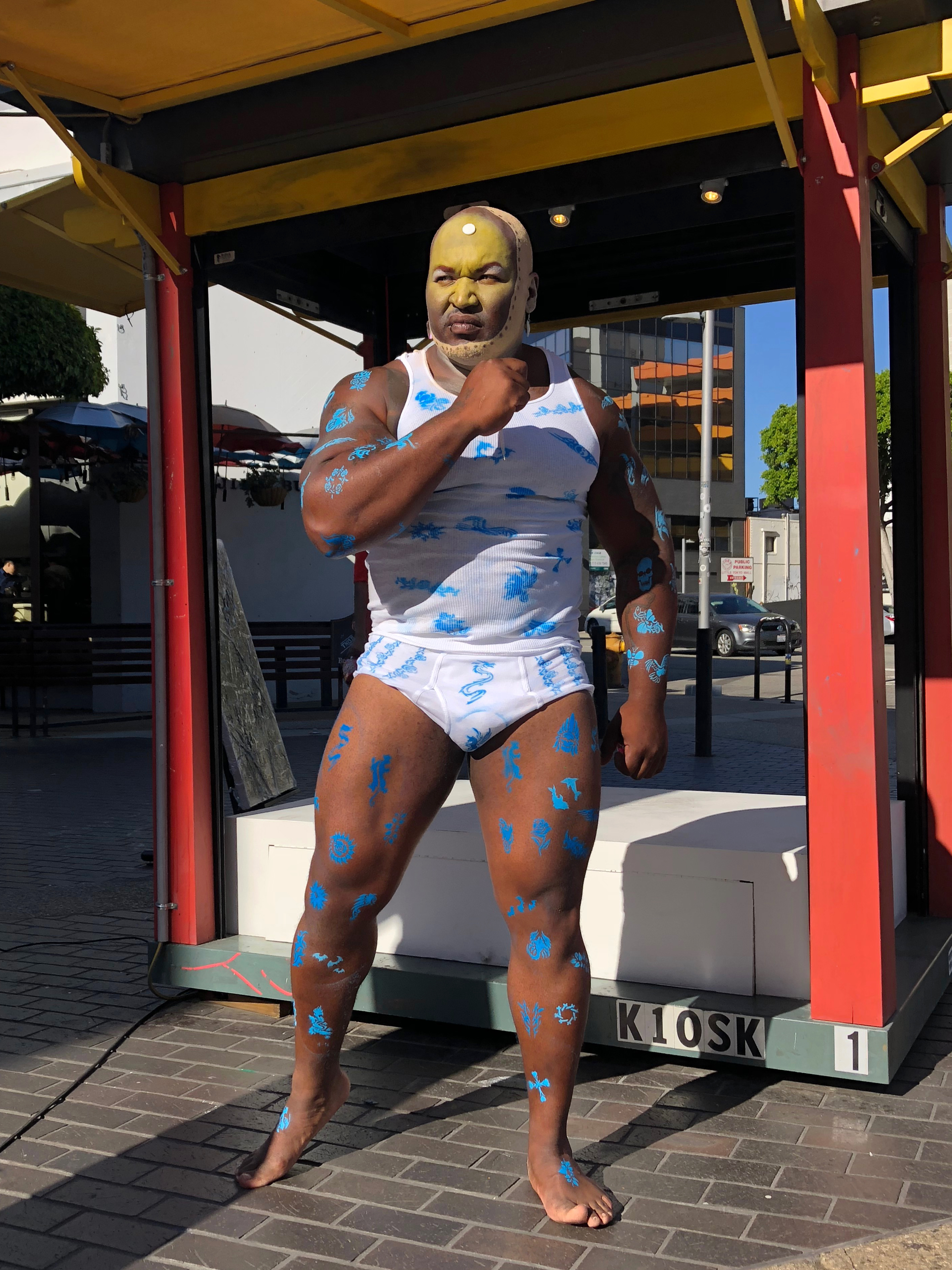 A muscular black man in tank top and underwear flexes outside in the daylight